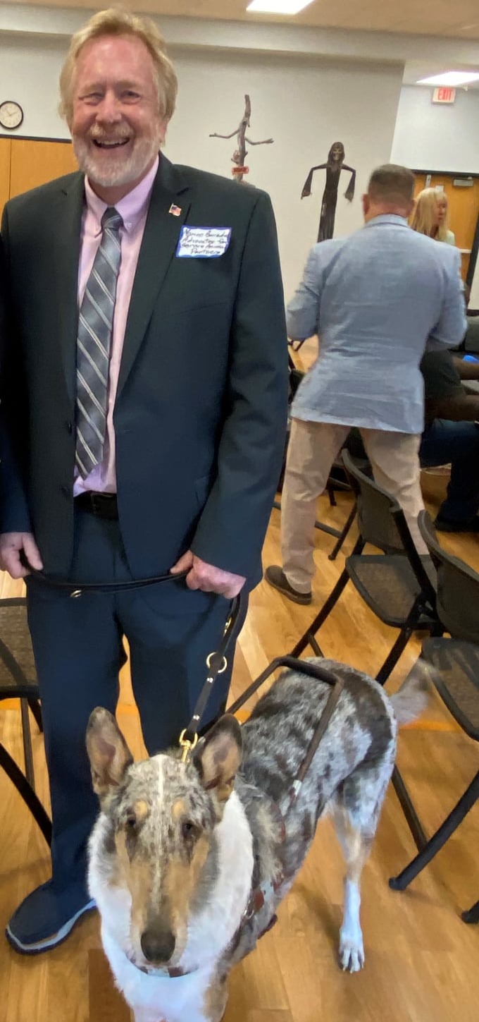 A white man with short graying red hair, smiling, stands in a community center with his smooth coat, Blue Merle Collie in harness by his side. 